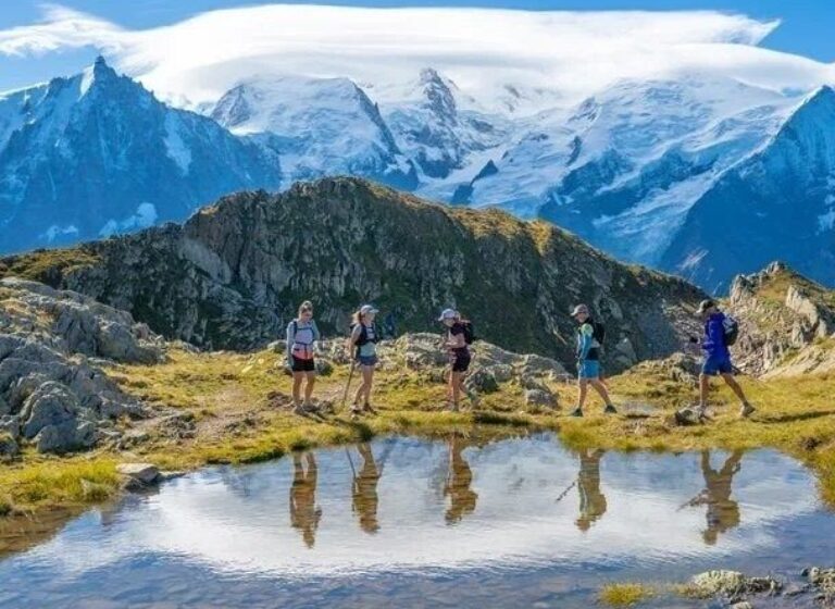 Trail runners cross a rocky alpine ridge beside a reflective pool