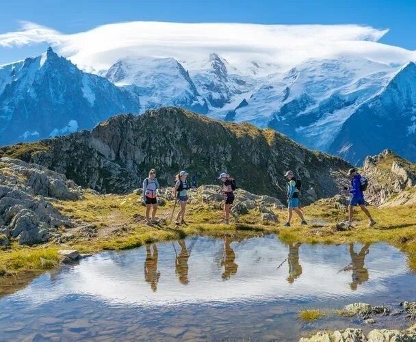 Trail runners cross a rocky alpine ridge beside a reflective pool